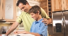 Photograph of a man with his arm on a boy's shoulder as the boy does homework