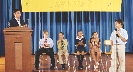 Photograph of several students seated onstage with one student at a microphone