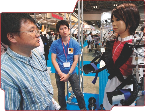 Photograph of two men looking at a robot the looks like a woman