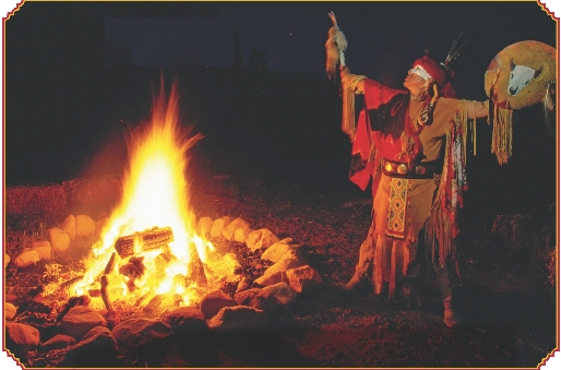 Photograph of a Native American by a fire at night in traditional clothing singing