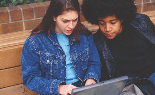 Photograph of a teenage girl using a laptop as a teenage boy looks on