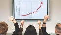 Photograph of cheering people in front a large graph that shows a sharp increase