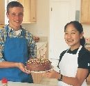 Photograph of a teenage boy and girl with aprons holding a cake together