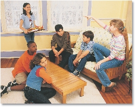 Photograph of same students as previous photo sitting around a coffee table as a girl serves snacks