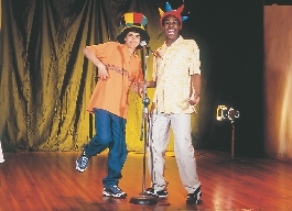 Photograph of two teen boys on stage—one wears an orange shirt and a large colorful hat, the other tan pants and a colorful crown