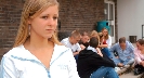 Photograph of a teenage girl standing by herself with a group of other teenagers sitting together in the background.