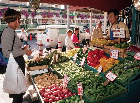 Photograph of a woman pointing to a particular vegetable in an outdoor vegetable stand as a worker looks on.