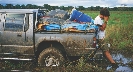 Photograph of a man with hand to his face and leaning against a truck with its wheels deep in mud