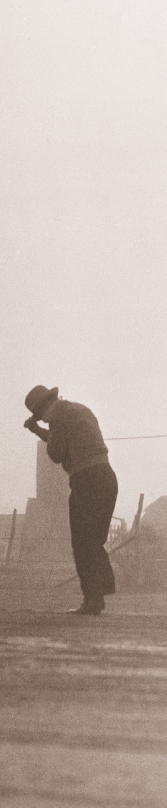 Photograph of a man holding onto his hat as wind blows dust (dry dirt) all around